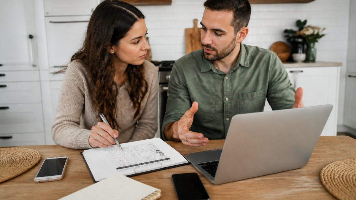 A couple sitting at a kitchen table with a laptop open to a vendor contact list, a printed checklist, two phones, and a notebook, mid-conversation while filling in vendor details