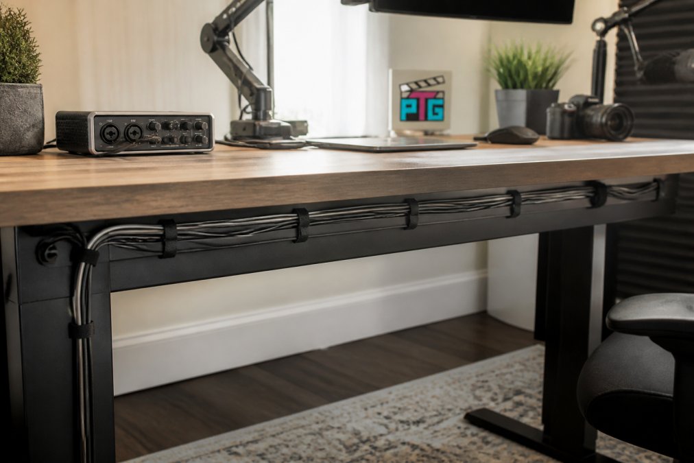 A home studio desk with cables neatly bundled in velcro ties and routed along the back edge, with a monitor arm and audio interface visible in a bright room