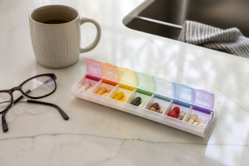 Close-up of a weekly pill organizer with compartments filled with different colored pills and capsules, sitting on a clean kitchen counter next to a coffee mug and reading glasses