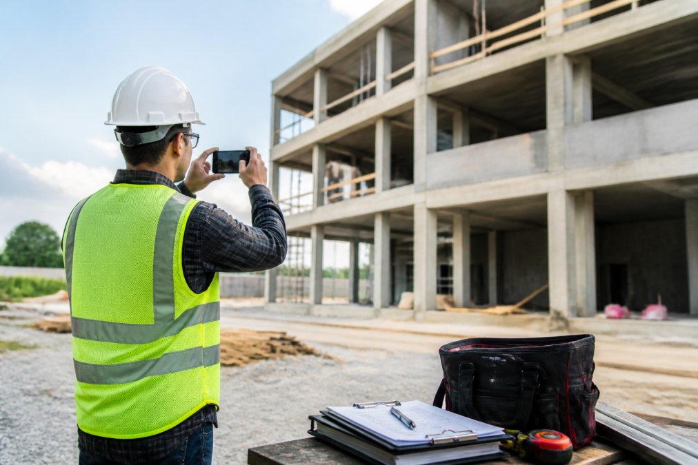 A field inspector in a hard hat and safety vest holding a smartphone, photographing a partially built commercial structure on a construction site, with a clipboard and tool bag nearby