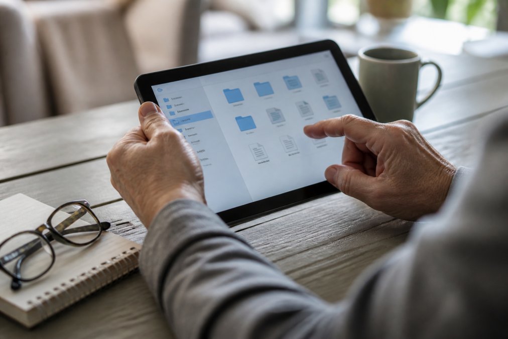 Close-up of hands using a tablet to access a secure digital filing system interface, with a notebook and reading glasses nearby on a desk