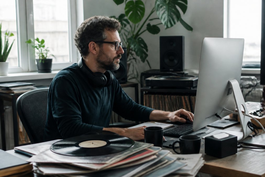 Music label manager reviewing artist roster data on computer screen in modern studio office, with headphones and vinyl records on desk, natural window lighting