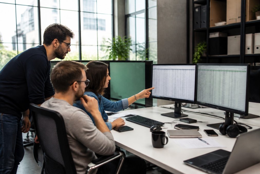 Label team collaborating around desk analyzing streaming metrics and release performance on multiple monitors, modern office setting with natural lighting