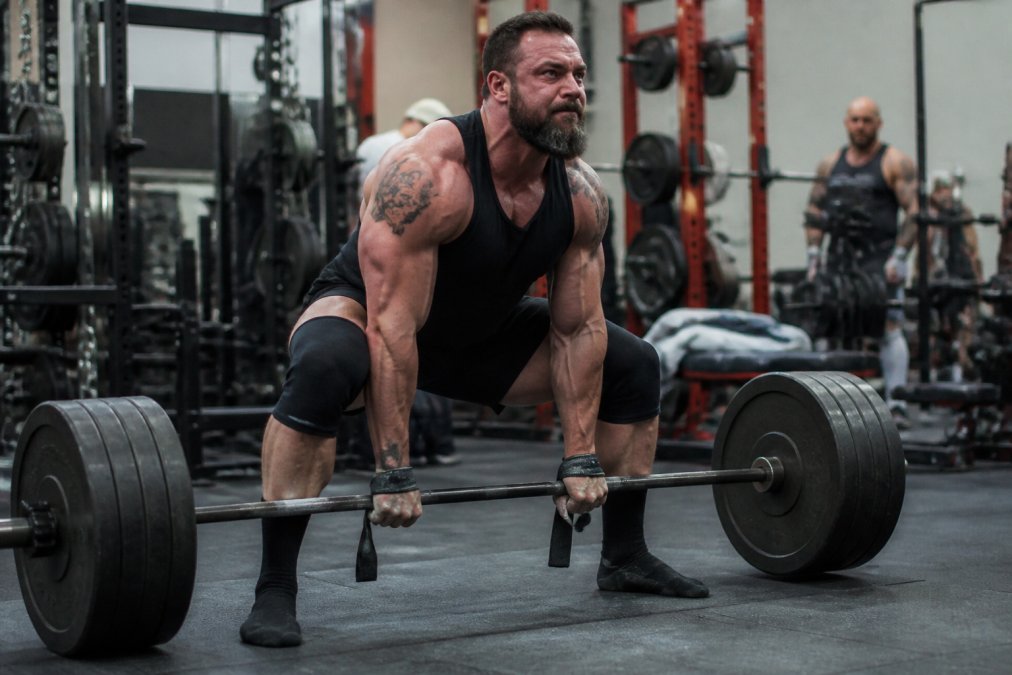 A focused lifter in a gym performing a heavy barbell deadlift, with visible strain and determination, wearing lifting straps and knee sleeves, surrounded by weights and gym equipment in a realistic training environment