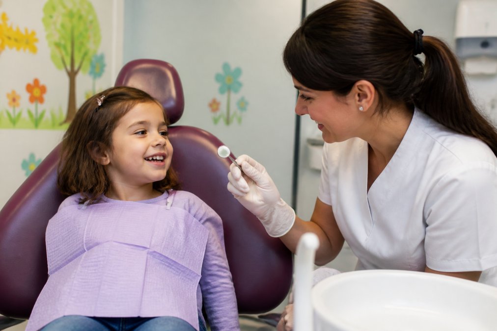 A young child sitting in a dental chair looking relaxed and smiling, with a gentle dentist in a white uniform showing them a small dental mirror, in a bright and welcoming practice with colourful wall art