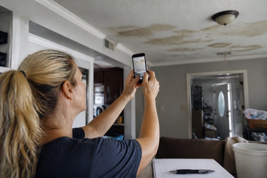 A homeowner using a smartphone to photograph water stains on a ceiling and walls, with a notepad and pen nearby for documenting damage details