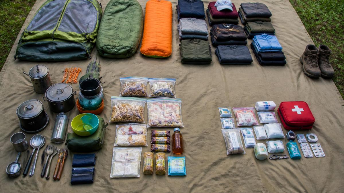 Overhead view of camping gear organized into distinct piles by category: tent with sleeping bag, folded clothing in neat stacks, cooking equipment and food items, and first-aid supplies arranged on a large canvas tarp in bright daylight
