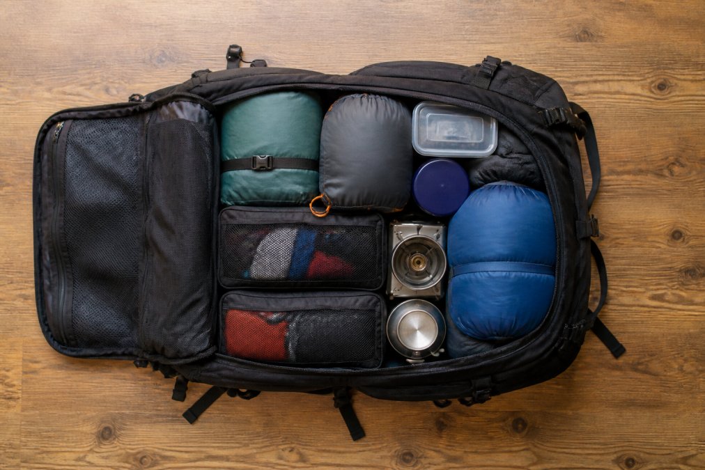 Overhead view of a large waterproof camping backpack laid flat with organized packing cubes, labeled storage containers, sleeping bag, tent, and cooking equipment arranged systematically inside on a wooden floor