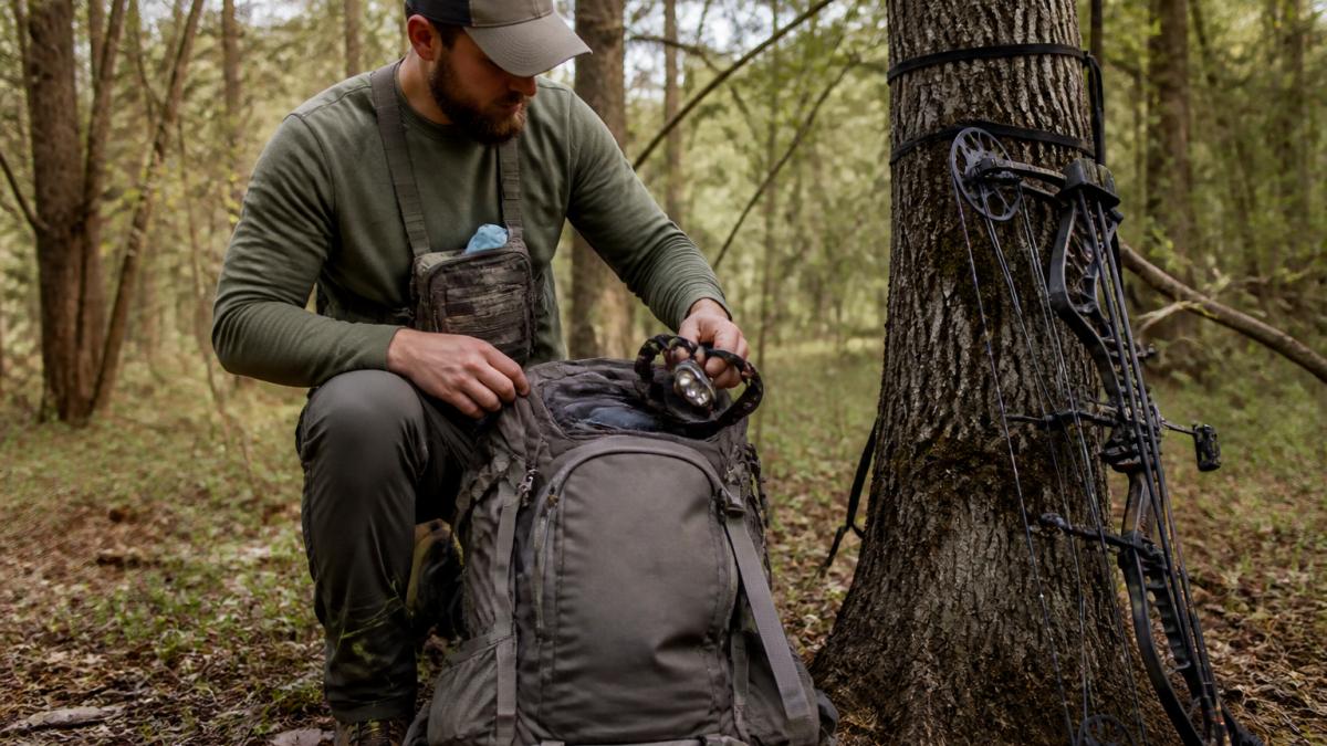 A backcountry hunter kneeling beside a fully loaded hunting backpack on a forest floor, pulling a headlamp from an open top pocket while a bow rests against a tree nearby