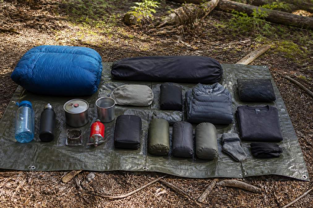 Backpacker's ultralight gear laid out on a tent footprint in natural outdoor setting, showing organized items including sleeping quilt, bivy sack, lightweight cookware, compression sacks, and moisture-wicking clothing arranged on forest floor with morning light