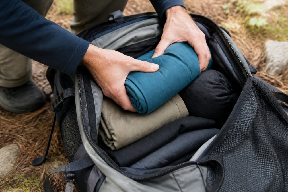Close-up of hands demonstrating roll-packing method with technical moisture-wicking fabric clothing inside open backpack, showing compression technique and efficient space usage in natural daylight