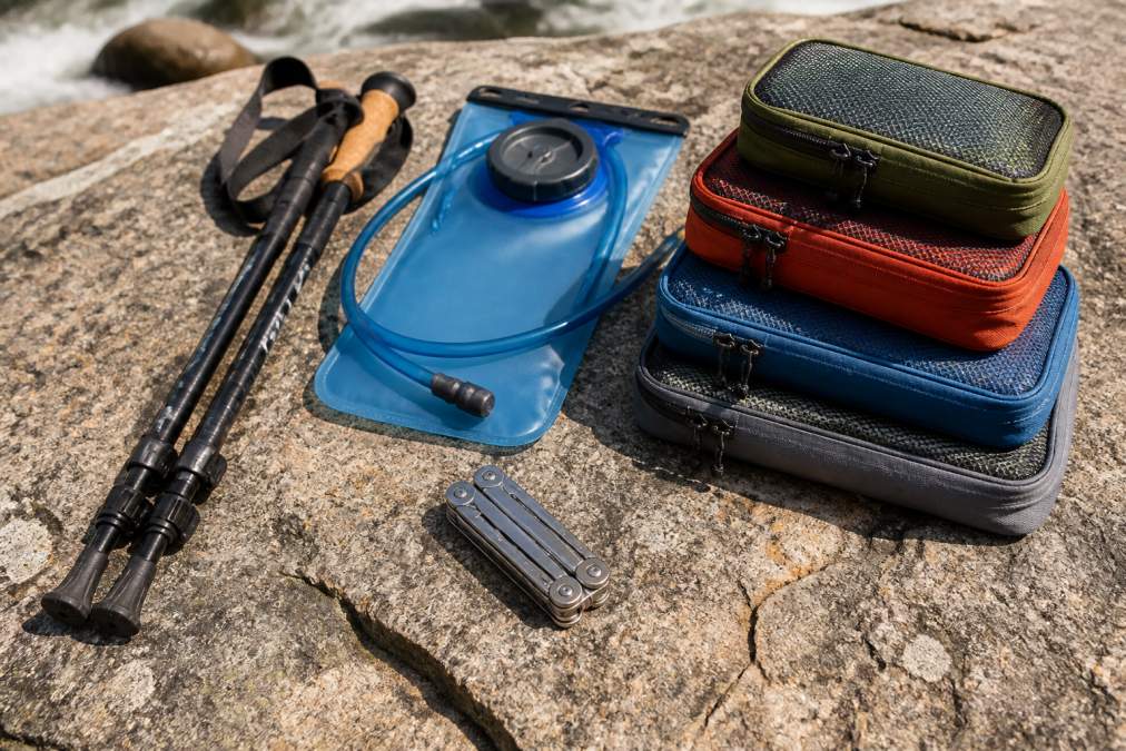 Close-up flat lay of lightweight hiking accessories including collapsible trekking poles, a compact hydration bladder, color-coded packing cubes, and a small multi-tool arranged on a textured natural stone surface in bright outdoor light