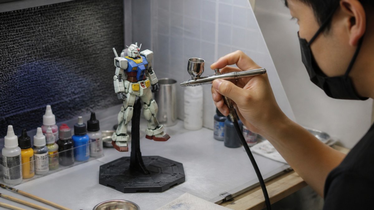 Hobbyist using an airbrush to paint a Gundam model on a stand, showing how to paint and customize gundam models.