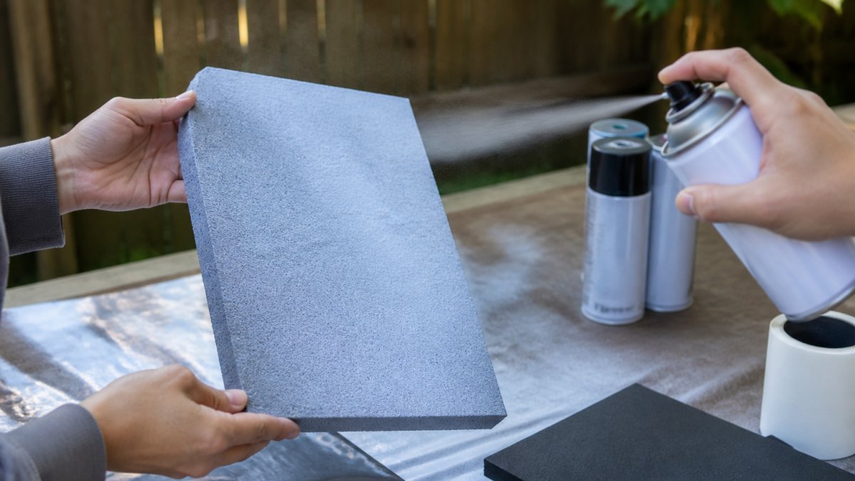Close-up of hands holding a craft foam sheet outdoors while spray paint is applied in light, even coats from proper distance away