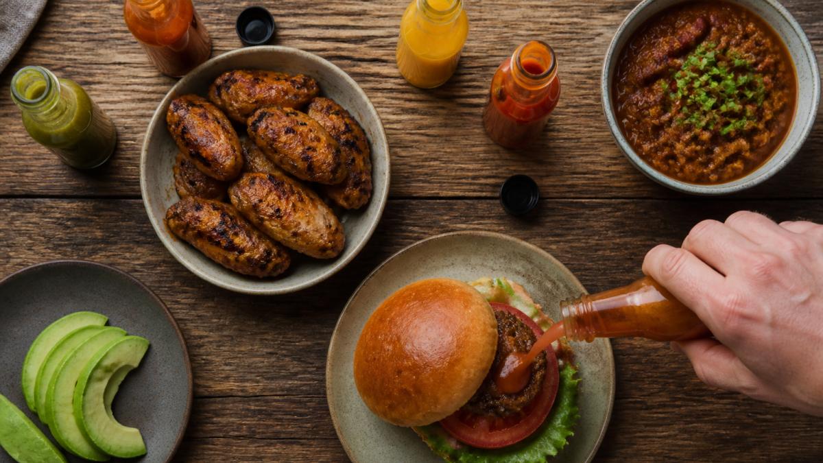 Overhead shot of a wooden table with grilled chicken wings, a burger, sliced avocado, a bowl of chili, and several open bottles of hot sauce, with a hand drizzling sauce over one dish