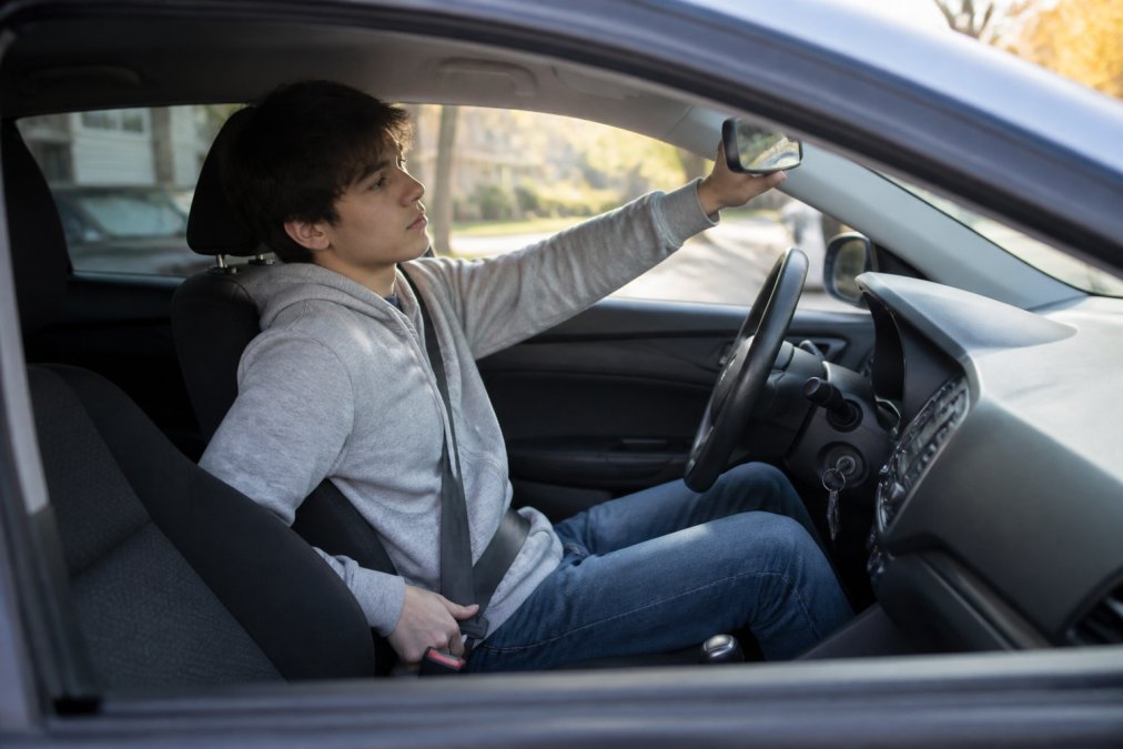 Young driver adjusting side mirror and checking seat position before starting vehicle in morning daylight, focused expression