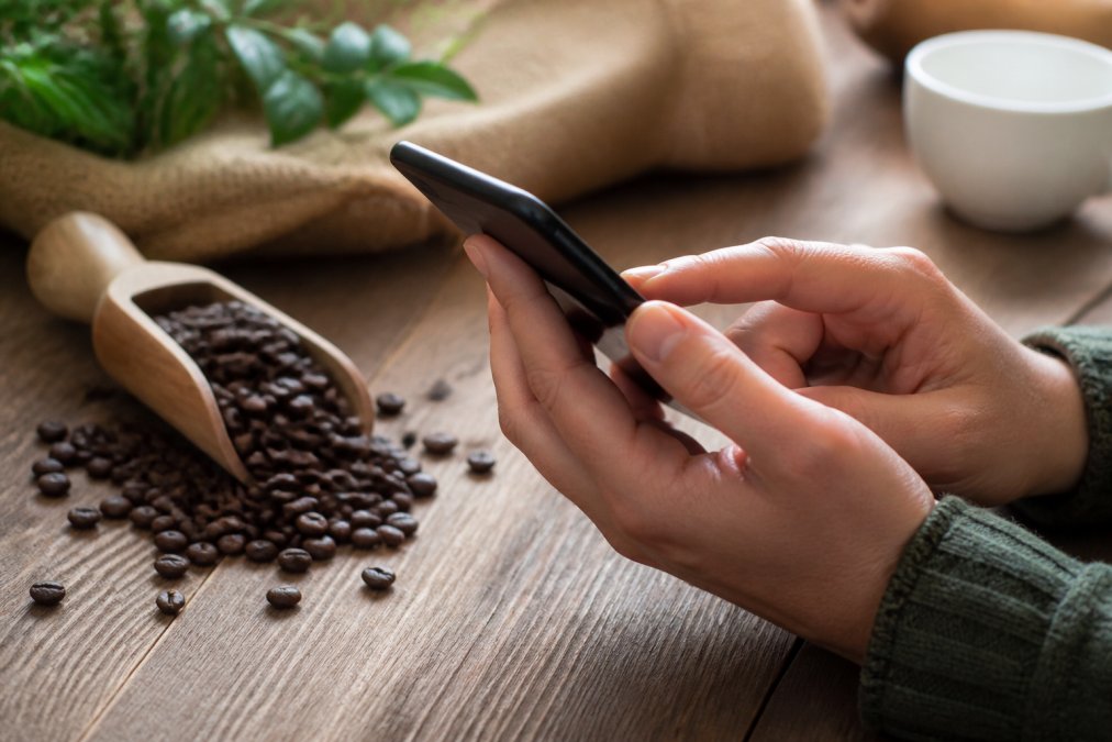 Close-up of hands using a smartphone to navigate a subscription management app interface, with fresh roasted coffee beans visible in a wooden scoop in the background