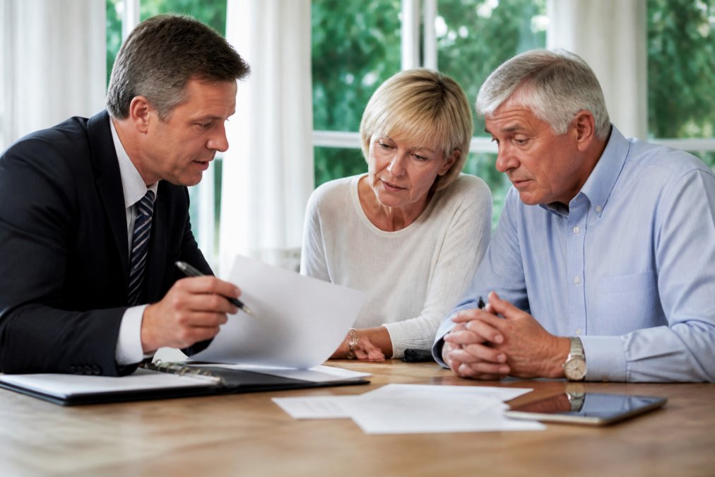 A financial advisor reviewing retirement paperwork with a couple at a bright desk, documents and a laptop open between them