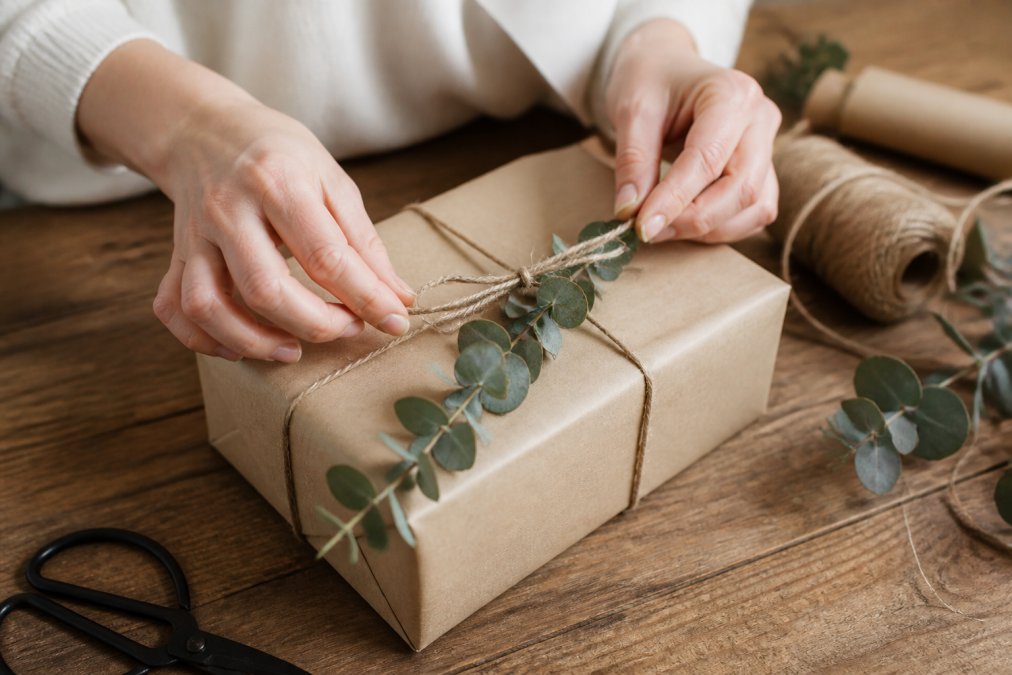 Hands carefully wrapping a gift with kraft paper and natural twine, a single eucalyptus sprig placed on top, refined wrapping technique on a wooden table with soft natural lighting