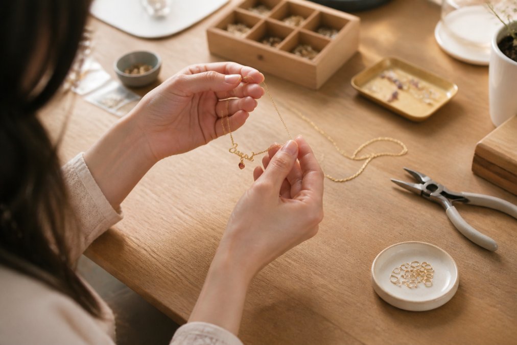 Close-up of a woman's hands holding a delicate gold name necklace with a small birthstone charm, soft natural light, warm and sentimental mood
