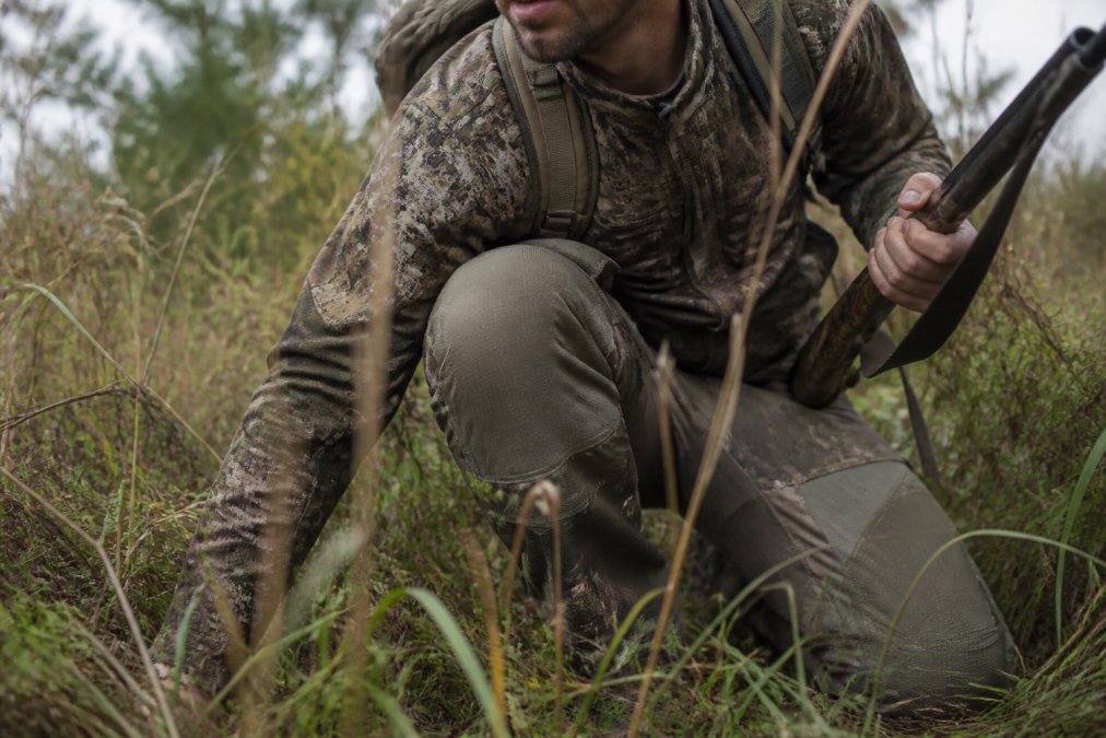 Hunter in camouflage crouching low in tall brush and grass, showing reinforced knees and articulated fit of durable hunting pants during active field movement