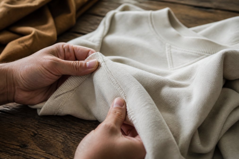 Close-up of hands examining the stitching and fabric of a high-quality cotton sweatshirt on a wooden table, natural light, cozy setting