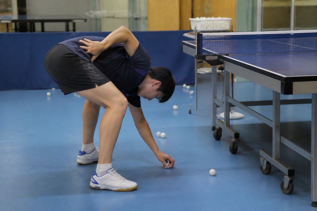A table tennis player bending over at the waist to pick up a white ball from the floor by hand, one hand pressed on the lower back, several balls scattered around a practice table