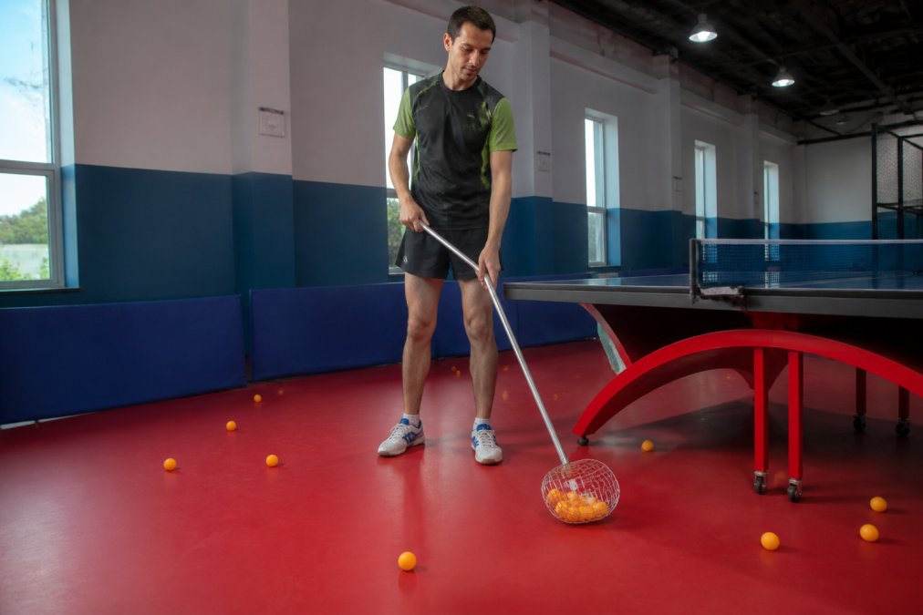 A table tennis player standing upright at a ping pong table, holding a long-handled ball retriever tool, with several orange ping pong balls scattered on the floor around the table in a bright practice room