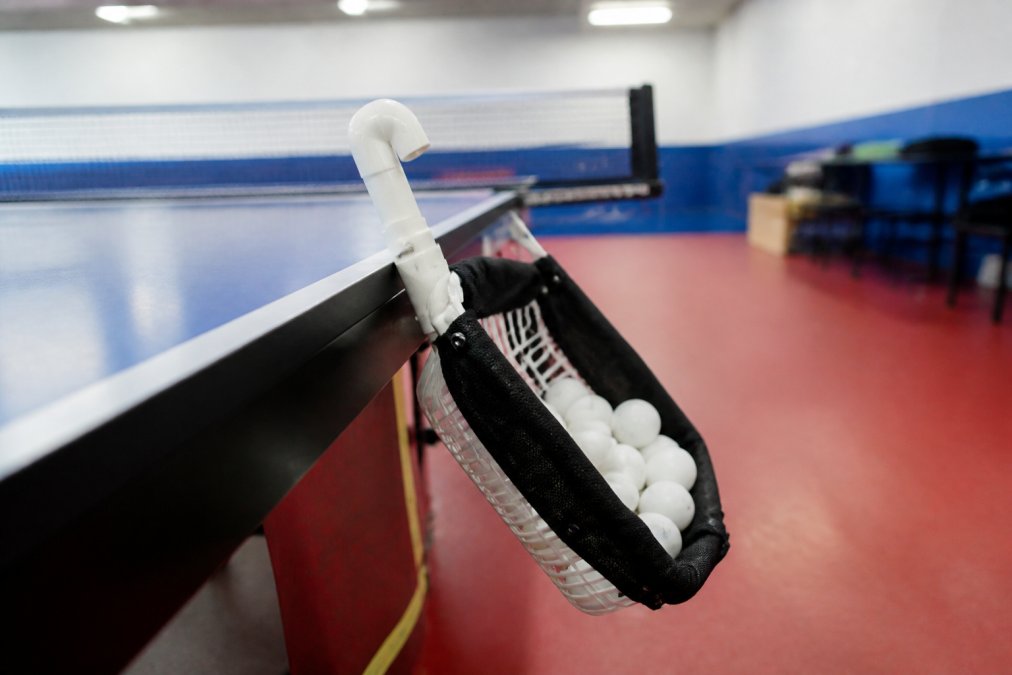 Close-up of a handmade table tennis ball picker tool resting against a ping pong table, with a small pile of white ping pong balls collected inside its basket or tube, in a well-lit practice room with overhead fluorescent lighting