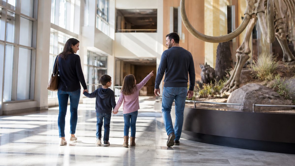A family with two young children walking hand-in-hand through a bright downtown museum lobby, pointing at a large interactive exhibit, with natural light streaming through tall windows