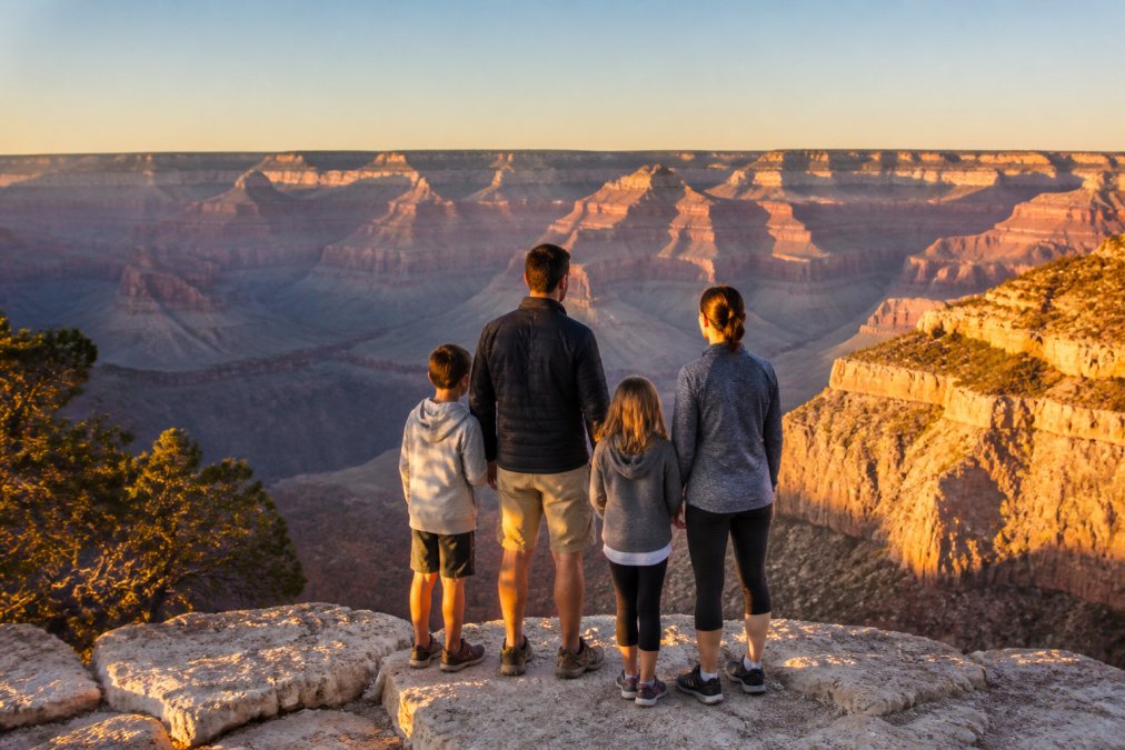 A family standing at a scenic overlook at the Grand Canyon South Rim during golden hour, layered canyon walls visible in the background under clear skies