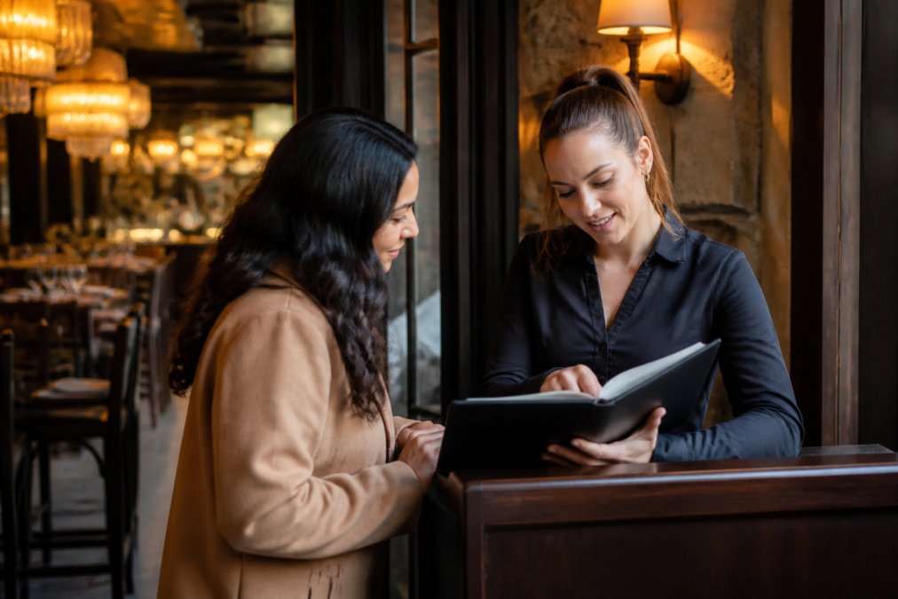 Restaurant host or manager reviewing a reservation book or tablet with a guest, discussing group booking details at the host stand, professional setting