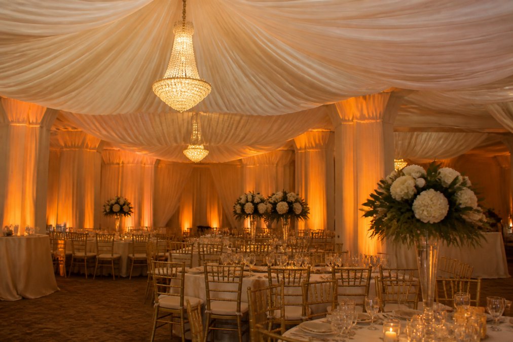 Elegant ballroom with luxurious ivory fabric draping cascading from the ceiling, illuminated by warm amber up-lights that accentuate the fabric folds and architectural columns