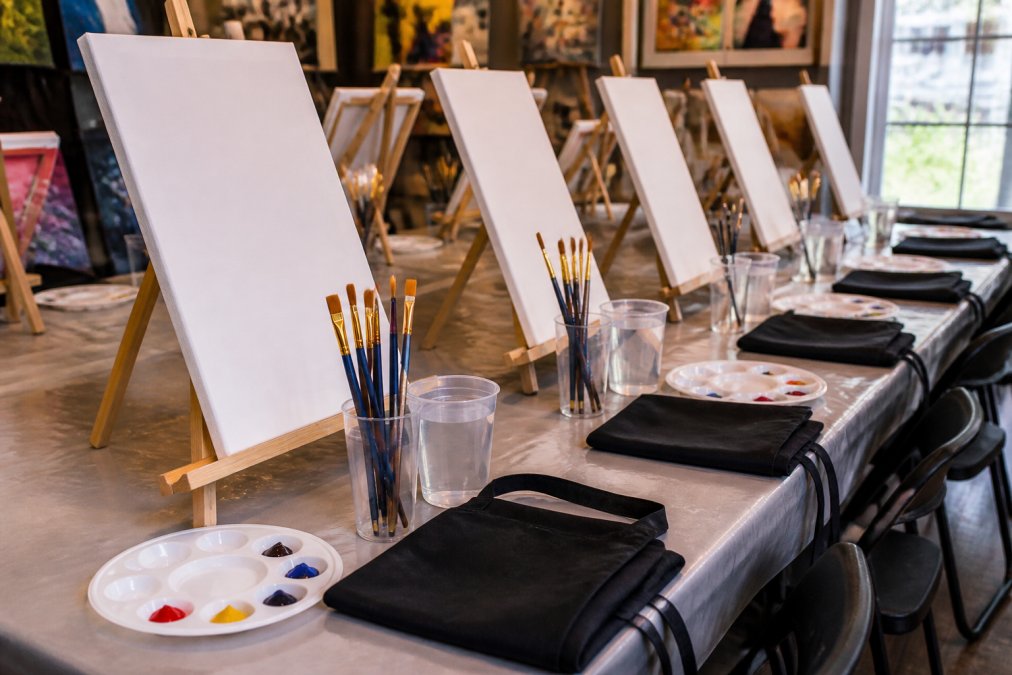 A paint and sip class table set up with canvases on easels, acrylic paint cups, brushes, palettes, water cups, and aprons ready for guests in a bright studio