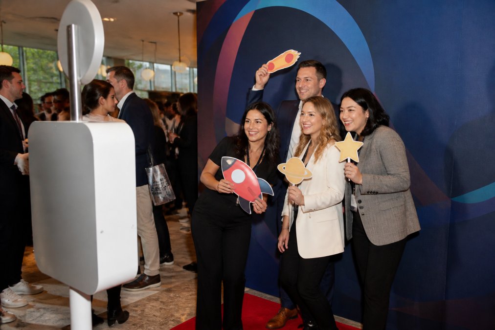 Professional photo booth setup with branded backdrop and custom props at a corporate product launch, attendees smiling and interacting with the booth, natural event lighting