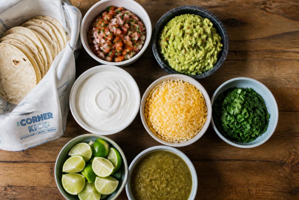 Overhead flat-lay of a fully loaded taco bar spread showing small bowls of pico de gallo, guacamole, sour cream, shredded cheese, cilantro, lime wedges, and salsa verde arranged on a wooden table with warm tortillas in a cloth-lined basket