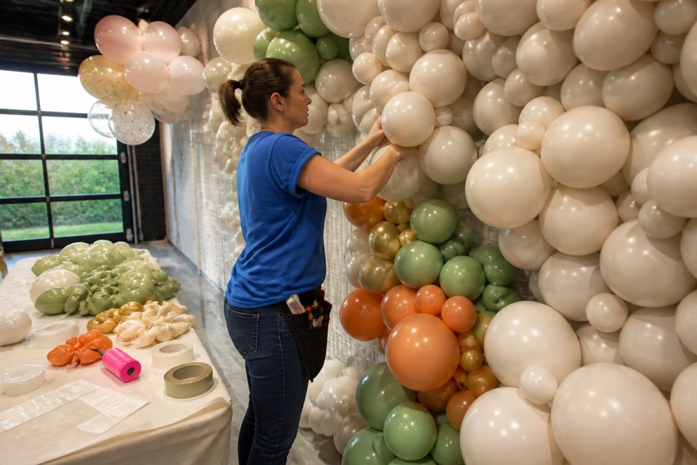Professional balloon artist at work installing a custom balloon backdrop for a kids party, carefully using glue dots and balloon tape to secure balloons to a wall in a bright venue space