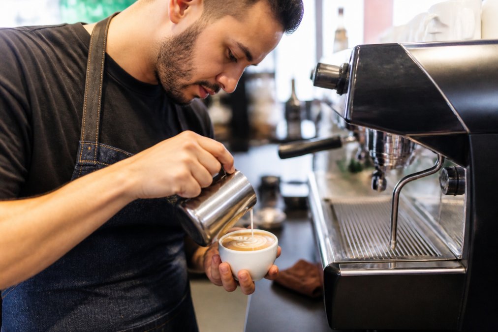 Skilled barista with focused concentration crafting latte art on specialty coffee drink, hands positioned precisely over espresso cup with milk steamer in background, professional espresso machine visible, warm café lighting