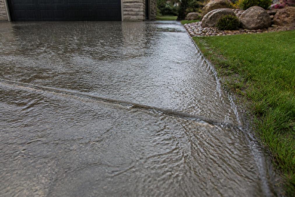 Close-up of water flowing down a sloped driveway surface during rain, demonstrating proper drainage angle with grass and landscaping visible at edges