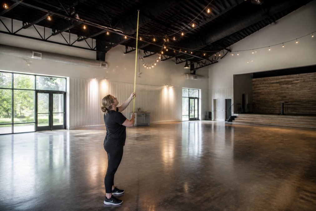 Professional balloon decorator standing in an empty event space, examining ceiling height and taking measurements with a tape measure, natural daylight from windows