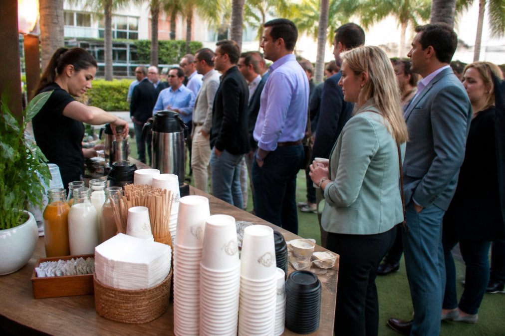 Busy beverage station with multiple guests waiting in line at a South Florida event, condiment station visible with various milk alternatives, cups, and napkins organized clearly
