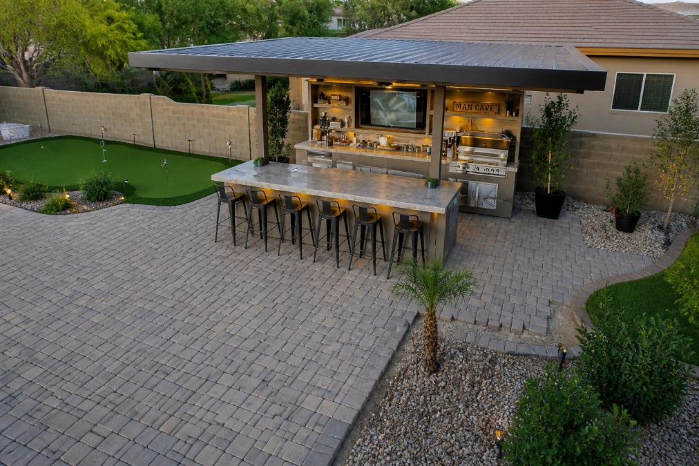 Overhead view of a backyard showing a completed L-shaped outdoor kitchen island with stainless steel grill, granite countertop workspace, and integrated seating area on a natural stone patio with landscaping