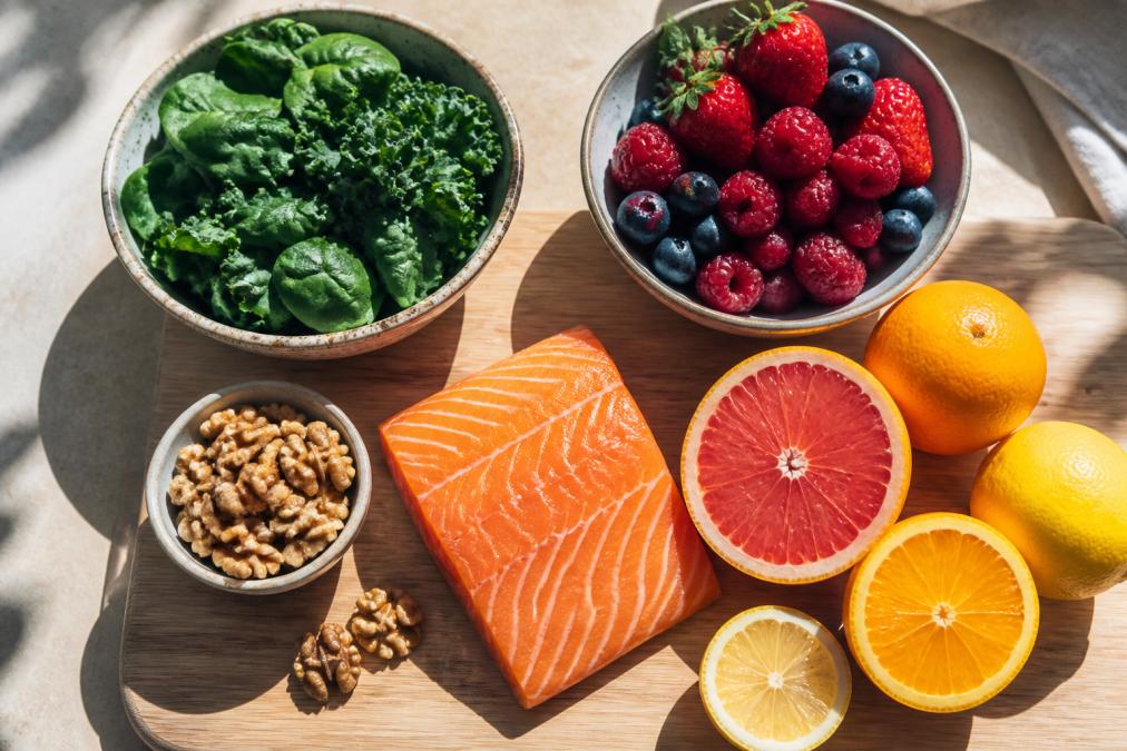Overhead shot of fresh salmon fillet, mixed berries, dark leafy greens, walnuts, and citrus fruits arranged on a light wooden board with natural morning sunlight casting soft shadows