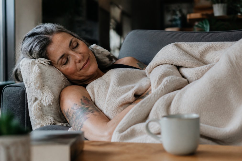 A woman in her 40s lying on a couch under a soft blanket with a cup of tea nearby, eyes closed, in a warmly lit living room, looking genuinely rested rather than posed