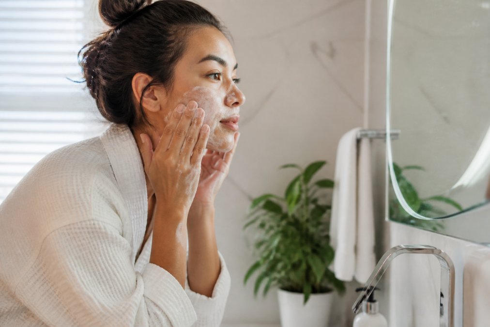 Woman applying cleansing product to her face with fingertips, standing at a bathroom sink with natural light, demonstrating proper cleansing technique