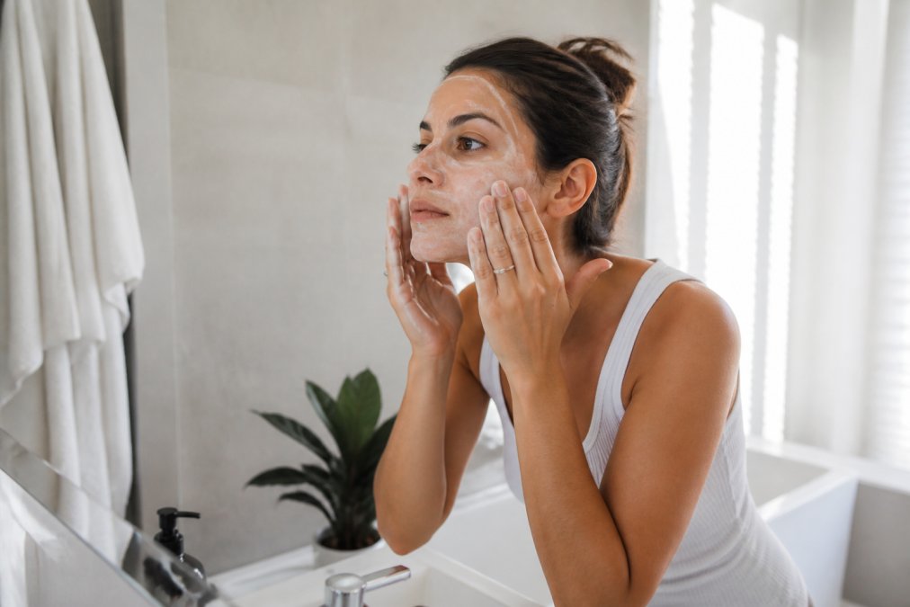 Person applying a gentle cleanser to face over a sink with soft morning light, showing proper technique with fingertips massaging in circular motions