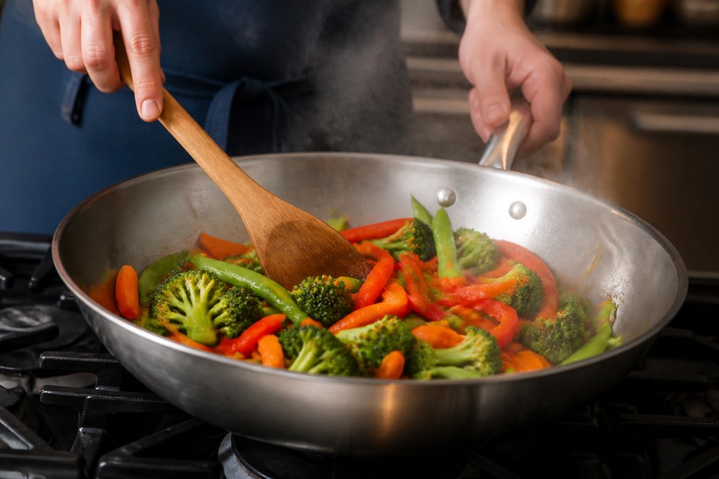 Close-up of hands quickly stir-frying colorful prepped vegetables (red peppers, broccoli, carrots, snap peas) in a hot stainless steel wok with visible steam rising, wooden spoon mid-stir, professional kitchen lighting