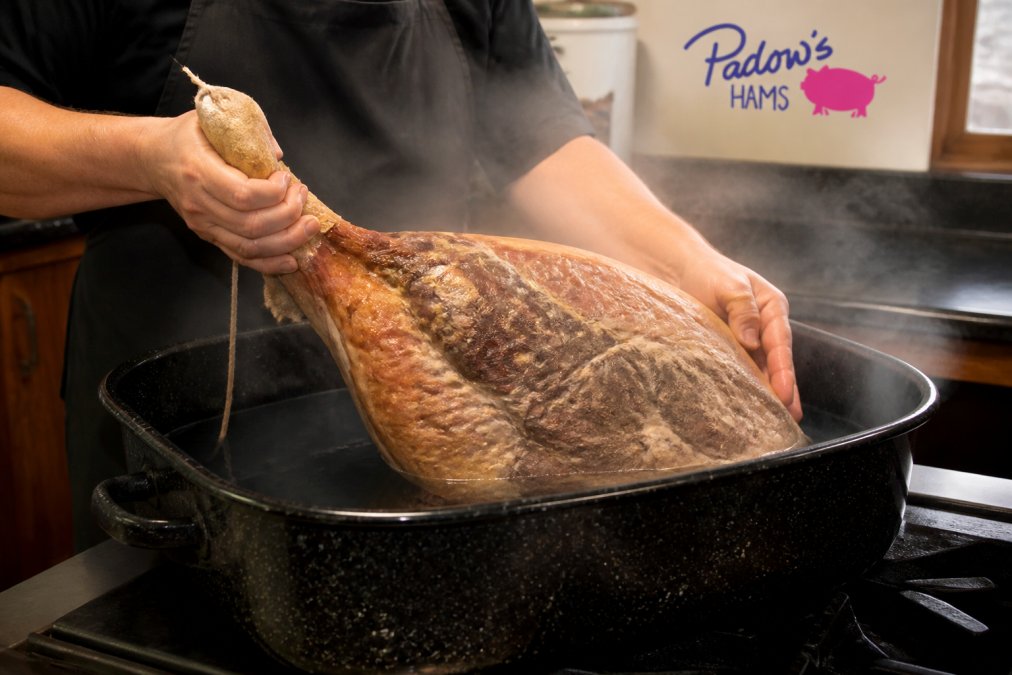 A home cook's hands lowering a whole salt-cured country ham into a large black speckled roasting pan filled with water, steam rising, warm kitchen lighting