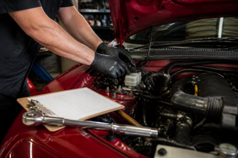 Close-up of a mechanic's hands in gloves checking the brake fluid reservoir under the open hood of a Mazda Miata in a well-lit garage, with a torque wrench and clipboard nearby