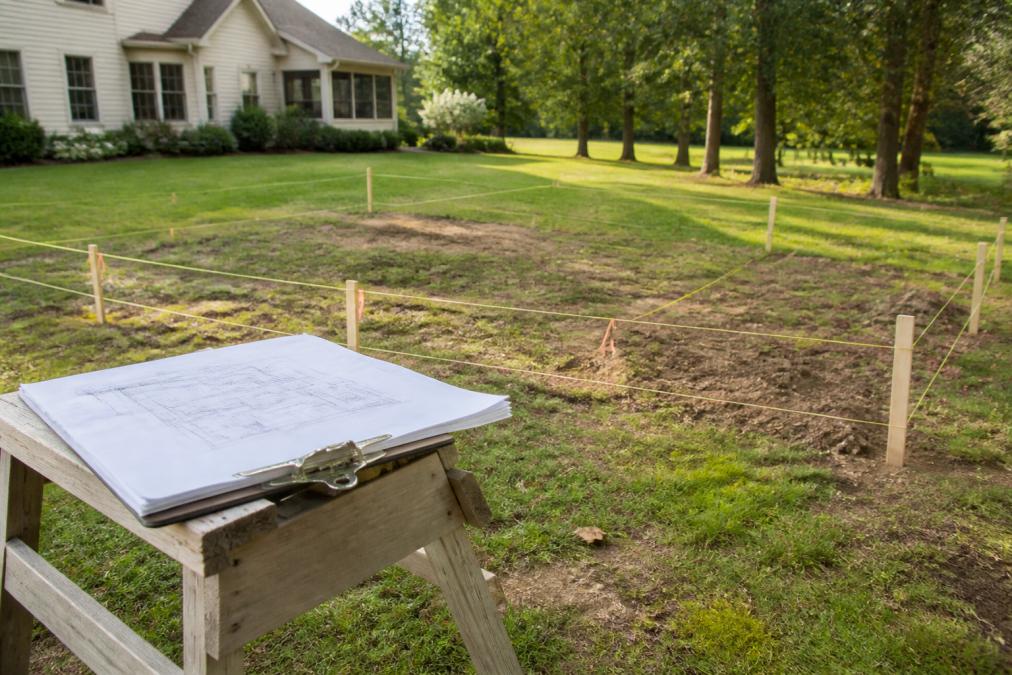 Residential backyard in Chesterfield with wooden stakes and string lines marking a future pool layout, a clipboard with site plans resting on a sawhorse, morning light